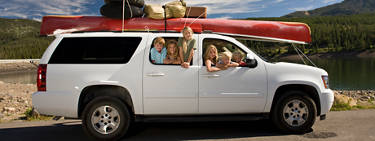 An image of a group of children in a white vehicle on a roadtrip looking out the windows with a long kayak on the roof.