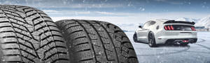 Two snow tires in front of a white Ford Mustang on snowy pavement