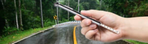 Hand holding a tire gauge against a wet road running through a forest